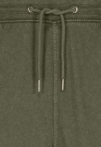 Gros plan sur un tissu vert olive avec un cordon de serrage noué et des œillets en métal, montrant les coutures détaillées et la texture.