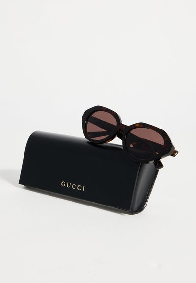 Lunettes de soleil rondes écaille avec des verres foncés reposant sur un étui noir Gucci avec des lettres dorées sur fond blanc.