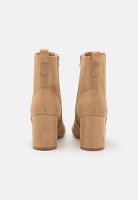 Botas de tobillo de ante marrón claro con un tacón block, con cierre de cremallera en la parte trasera y detalle de asa en bucle. Textura suave y diseño sencillo.