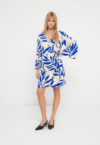 Omslagskjole i beige med blått løvmønster, med lange ermer, belte i midjen og kort skjørt. Kombinert med svarte spisse hæler.