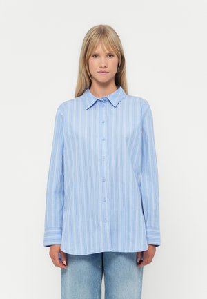 Žena nosí světle modrou košili s dlouhým rukávem s bílými svislými pruhy a světlé modré džíny, stojící na jednoduchém pozadí.