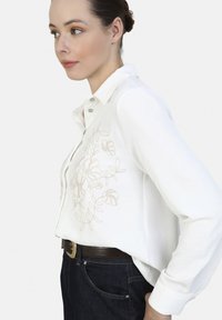 White button-up shirt with long sleeves, featuring beige floral embroidery at the chest, paired with dark denim jeans and a brown belt.