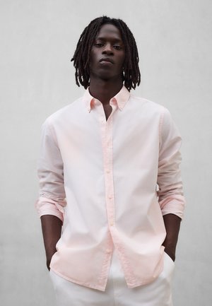 Mann mit Dreadlocks, der ein hellrosa Hemd mit hochgekrempelten Ärmeln trägt und mit den Händen in den Taschen einer weißen Hose steht.