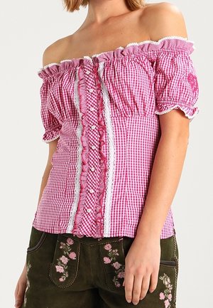 Kvinde iført en lyserød og hvid ternet bluse med bare skuldre, blonder og knapper, sammen med mørkegrønne bukser broderet med lyserøde blomster.