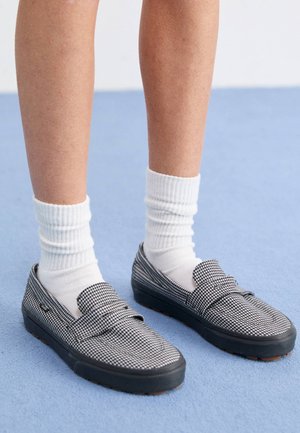 Benedenbenen met witte ribgebreide sokken en zwart-witte loafers met pied-de-poule patroon op een lichtblauwe vloer.