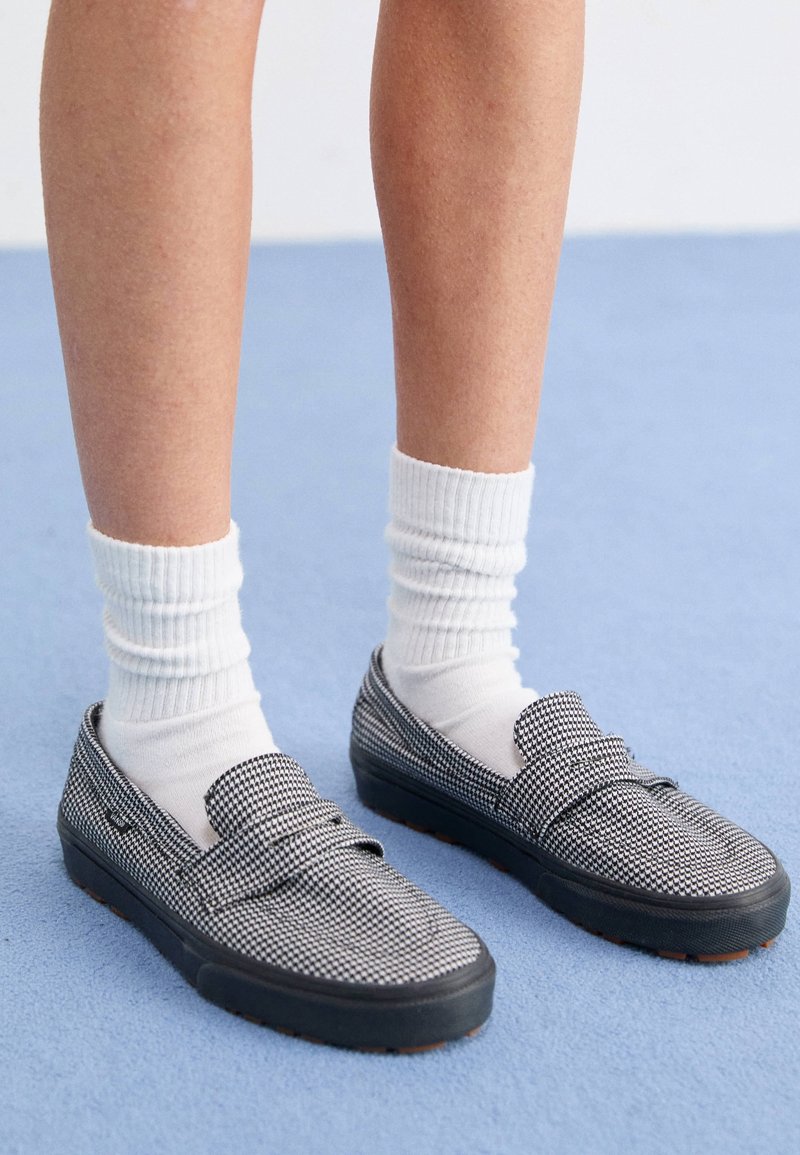 Jambes portant des chaussettes blanches côtelées et des mocassins slip-on pied-de-poule noir et blanc sur un sol bleu clair.