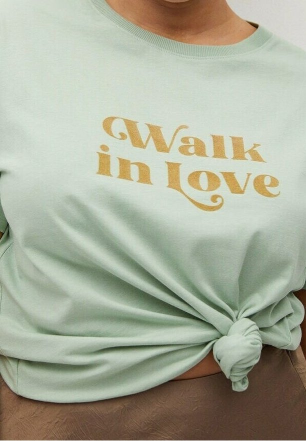 Chemise vert clair nouée à la taille avec un texte doré « Walk in love » sur la poitrine.
