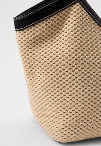 Beige woven tote bag with a textured finish and a black leather top rim, featuring white stitching along the edge.