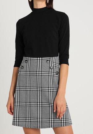 Haut noir moulant à col montant, associé à une jupe à carreaux gris et noir avec deux gros boutons sur le côté.