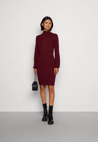 Vinröd ribbad stickad klänning med hög halsringning, figurnära silhuett och långa ärmar. Bärs med svarta ankelboots och en liten handväska.