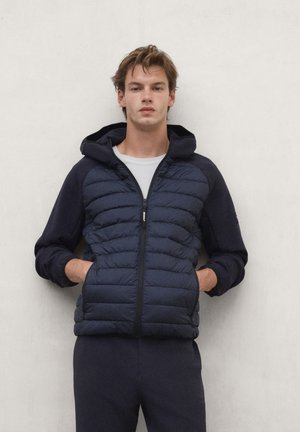Young man wearing a navy hooded quilted jacket over a white shirt with hands in pockets, standing against a plain light wall.