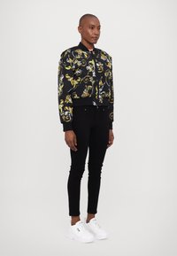 Cropped black bomber jacket with yellow and white swirling floral pattern, ribbed cuffs and hem, paired with black skinny jeans and white sneakers.