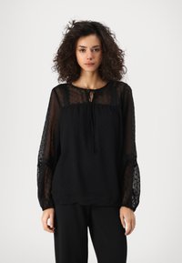 Blusa nera con maniche a pois trasparenti, scollo rotondo con dettaglio a fiocco e vestibilità morbida. Il tessuto testurizzato aggiunge un interesse visivo.