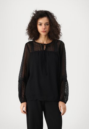 Black blouse featuring sheer dotted sleeves, a round neckline with tie detail, and a relaxed fit. Textured fabric adds visual interest.