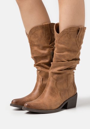 Bottes en cuir brun mi-mollet avec talons carrés bas portées sur jambes nues, dotées de languettes de tirage et de détails de couture subtils.
