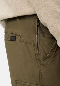 Nahaufnahme von olivgrünen Hosen mit seitlicher Reißverschlusstasche und Camel Active Markenetikett, getragen mit einem beigen Strukturpullover.