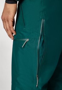 Tyrkysové softshellové nohavice s vreckami na zips, hladkou textúrou a štíhlym dizajnom, s kontrastnými zipsovými detailmi z pohľadu funkčnosti.