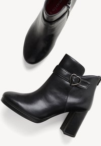 Botine negre din piele, cu bot curbat, toc înălțat și o cataramă decorativă în formă de inimă pe o curea în jurul gleznei.