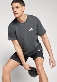 Black athletic t-shirt with textured fabric and a white Adidas logo on the chest, paired with black shorts, holding a black kettlebell.
