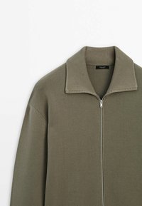 Pull en olive zippé avec un col et des poignets côtelés, fabriqué en tissu tricoté lisse. Il dispose d'une fermeture éclair argentée et d'une coupe décontractée.