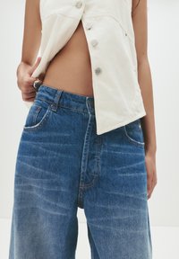Une personne portant un jean en denim bleu et un gilet beige déboutonné, la main sur la taille, se tenant devant un fond clair.