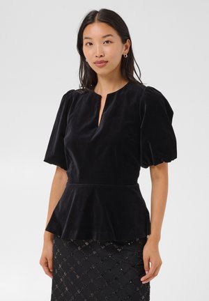 Femme aux longs cheveux foncés portant un chemisier en velours noir à manches bouffantes avec un décolleté en V et une jupe noire à motifs sur un fond uni.