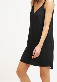 Černé šaty bez rukávů s výstřihem do V, vyrobené z lehkého materiálu. Mají volný střih a sahají nad kolena. Jednoduchý design, bez vzorů.