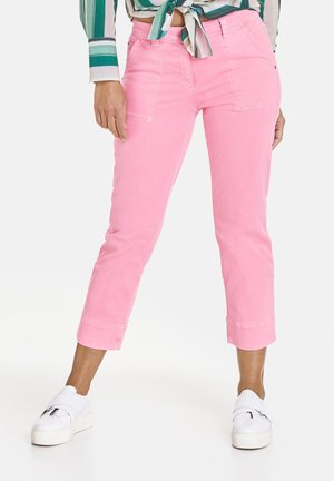 Femme portant un pantalon court rose, des baskets blanches et une chemise verte rayée nouée, debout, jambes écartées sur fond blanc.