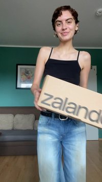Kardboonist Zalando kast, mida hoiab inimene mustas tanksärgis ja helesinistes laiemates teksades. Tuba, kus on rohelised seinad ja diivan.