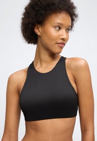 Žena s krátkymi kučeravými vlasmi v čiernom rebrovanom krátkom tielku bez rukávov, pozerajúca sa doprava na jednoduchom svetlom pozadí.