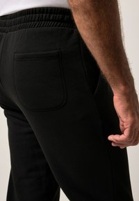 Close-up van de hand van een persoon die naast hun heup rust, met zwarte broek aan met een elastische tailleband en een achterzak.