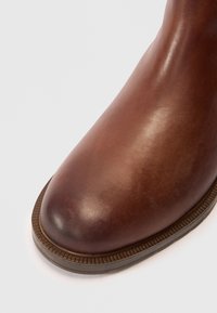 Bocanci din piele maro până la gleznă, cu o finisare netedă, vârf rotunjit și talpă din cauciuc texturată. Cusături distinctive pe margine.