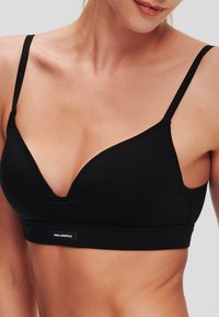 Bralette en coton noir avec un décolleté en V profond, des fines bretelles ajustables et une bande logo en bas. Texture lisse, style minimaliste.
