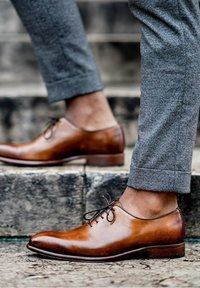 Des chaussures habillées en cuir marron avec une finition brillante, un bout rond et des lacets fins, associées à un pantalon à motifs gris, se tenant sur des marches en pierre.
