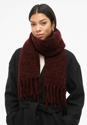 Sciarpa di maglia bordeaux con una texture morbida, caratterizzata da estremità frangianti. Indossata in modo sciolto attorno al collo su un cappotto nero, creando un effetto sovrapposto.