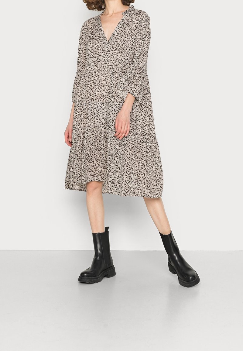 Robe à imprimé floral beige avec un décolleté en V profond, manches longues et jupe à volants. Associée à des bottines noires épaisses.