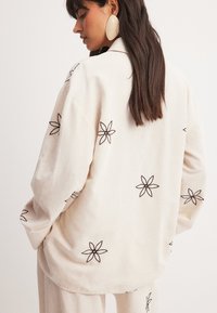 Béžový košový kabát vyrobený z ľanu, s čiernymi kvetinovými výšivkami, voľný strih, klopový golier a spadnuté ramená.