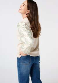 Sweatshirt beige avec des manches à motif floral irisé. Associé à un jean droit bleu. Texture lisse et coupe décontractée.