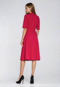 Fuksjowa sukienka z krótkimi rękawami, dopasowanym górnym partem i rozkloszowaną spódnicą. Tkanina wydaje się gładka. Noszona z czarnymi butami na wysokim obcasie.