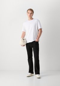Hvit bomulls t-skjorte med korte ermer og logo, kombinert med sorte jeans og hvite joggesko, holder en beige stoffpose. Enkelt design.