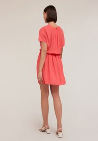 Korallenfarbenes kurzes Kleid mit kurzen Ärmeln, geraffter Taille und subtiler Rückenfalten. Kombiniert mit weißen, rückenfreien Schuhen. Glattes, leichtes Material.