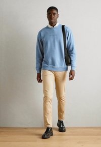 Lichtblauwe sweatshirt met geribde boorden, beige slim-fit broek en zwarte veterschoenen. Schoudertas over de schouder.