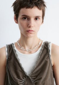 Silberne Kettenhalskette kombiniert mit einem geschichteten hellgrauen Tanktop und einem braunen Samtweste mit Spitze und Raffungen.