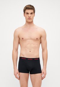 Bóxers negros ajustados con una cinturilla elástica roja que presenta la marca "HUGO". Material suave, diseño ajustado y longitud hasta medio muslo.