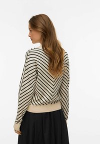 Maglione beige chiaro con strisce a chevron nere, polsini e orlo a coste, caratterizzato da una vestibilità ampia e una morbida texture. Abbinato a una gonna nera.