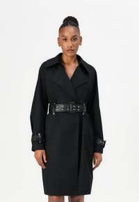 Zwarte trenchcoat met een brede kraag, ceintuur in de taille met oogjes en details op de manchetten. Glad materiaal met een rechte silhoette.