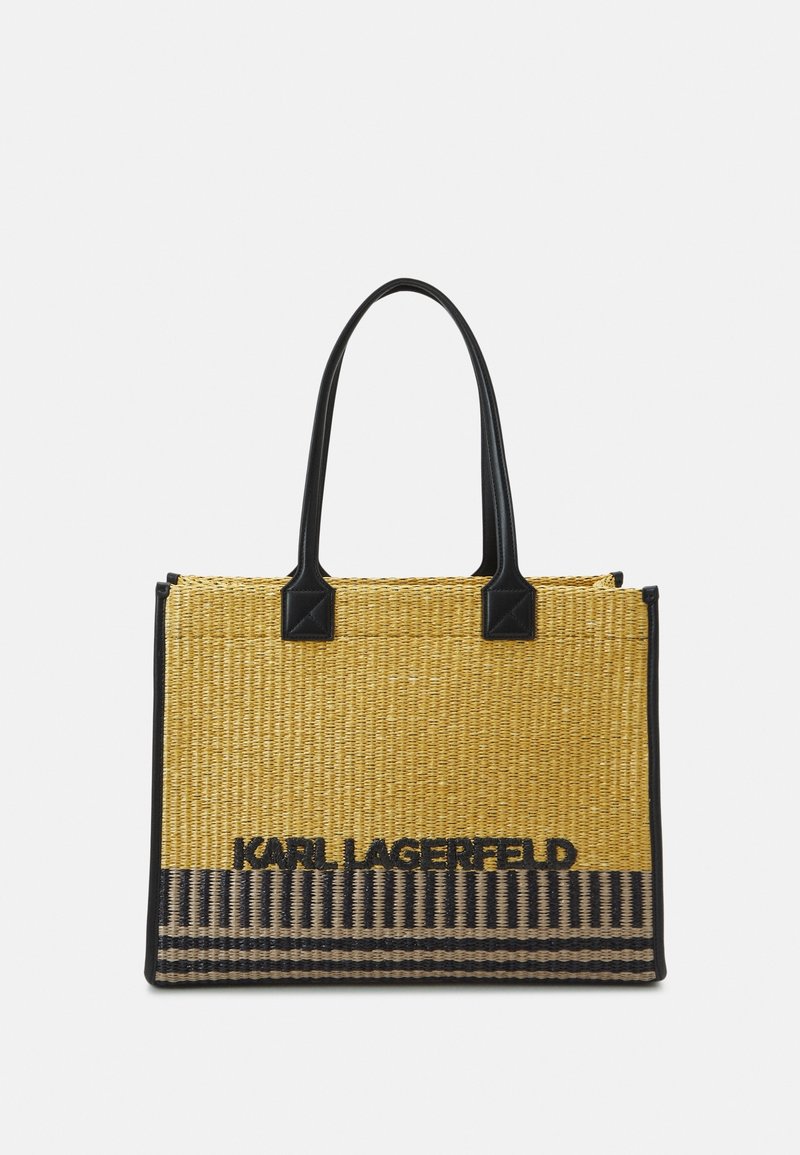 Bolso tote de paja tejido en amarillo con asas y detalles de cuero negro. Cuenta con el logo "Karl Lagerfeld" y un patrón de rayas en la parte inferior.