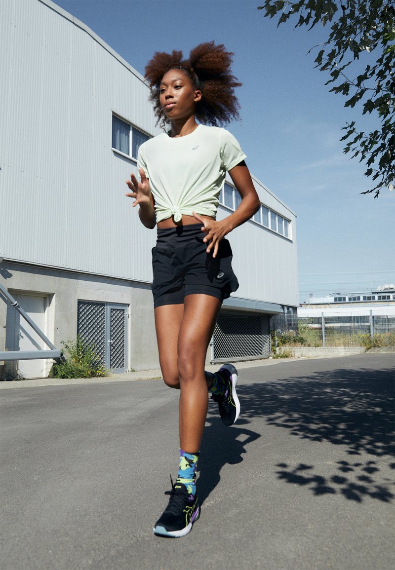 Modelo con una camiseta anudada verde claro y pantalones cortos negros, llevando calcetines de colores con estampados y zapatillas negras para correr, trotando por un sendero.