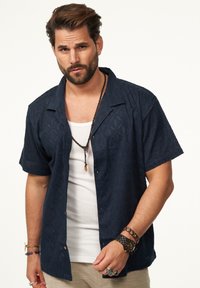 Marineblaue Kurzarmbluse mit strukturiertem Blumenmuster, offenem Kragen und Knöpfen. Über einem weißen Top getragen und mit Schmuck accessoriert.