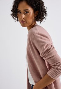 Un cardigan rose clair en texture douce, à col ouvert, avec des poignets côtelés, porté par-dessus un haut blanc. L'arrière-plan est neutre.